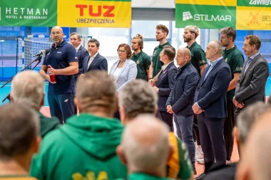 Mężczyzna w granatowym dresie przemawia do publiczności przez mikrofon na hali sportowej, za nim stoją oficjele i sportowcy w zielonych koszulkach pod banerami
